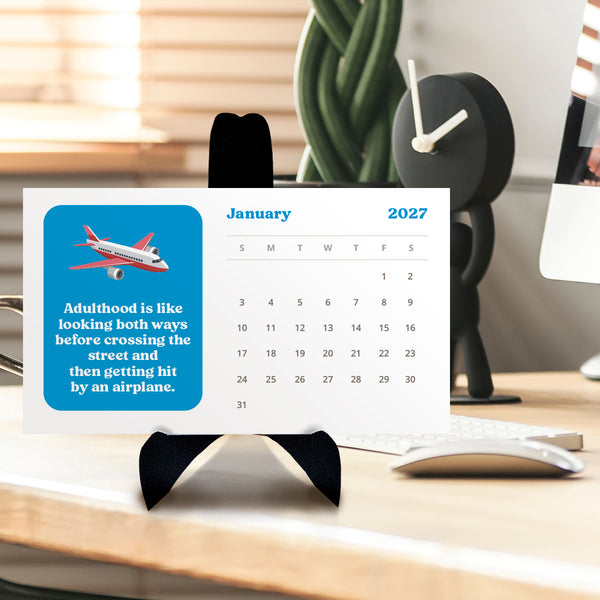 A monthly desk calendar for January 2027 displayed on a black stand on a wooden desk, with a blue graphic featuring an airplane and a quote in the foreground. A plant, a black desk clock, and a computer monitor are blurred in the background.