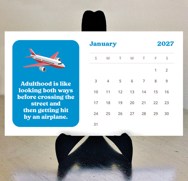 A 2027 desk calendar page displayed on a black stand against a light beige wall. The calendar has a white background with a bright blue box on the left containing an airplane illustration and a humorous quote about adulthood.