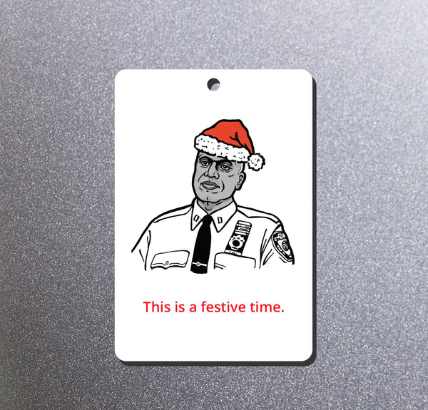 A rectangular white Christmas ornament hanging from a red ribbon, featuring a black-and-white illustration of Brooklyn Nine Nine's Captain Holt wearing a red Santa hat, with the red text 'This is a festive time.' photographed on a textured gray background.