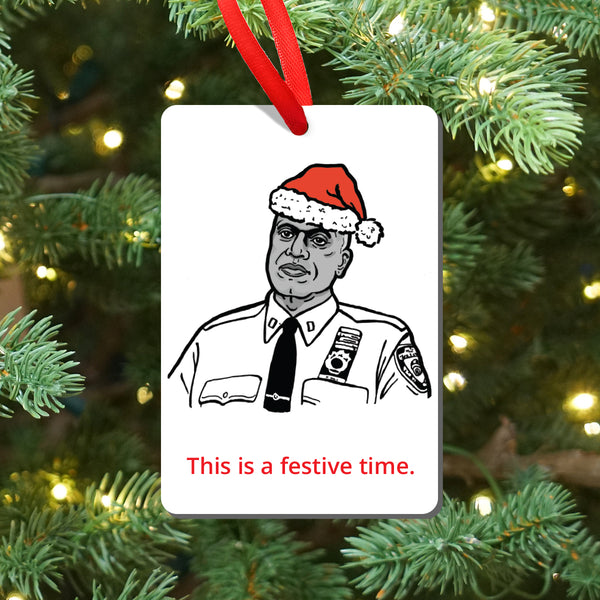 A rectangular white Christmas ornament hanging from a red ribbon, featuring a black-and-white illustration of Brooklyn Nine Nine's Captain Holt wearing a red Santa hat, with the red text 'This is a festive time.' The ornament is photographed against a blurred Christmas tree background with green branches and warm lights.