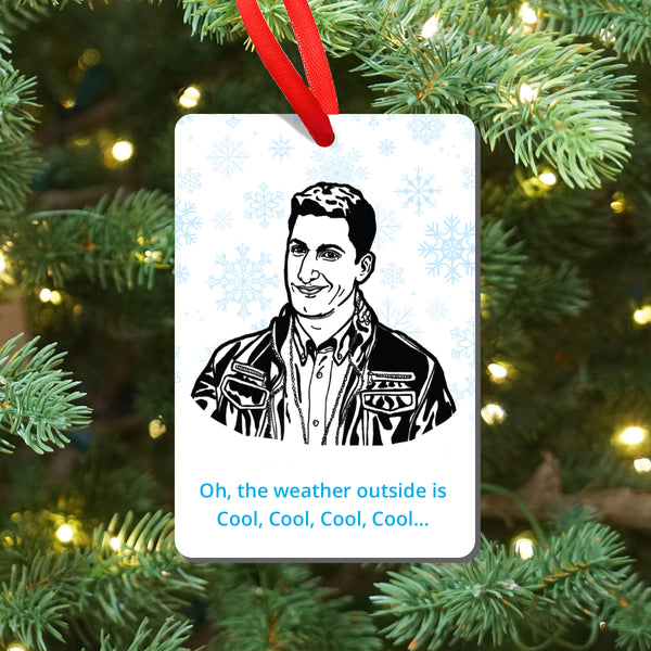 A Christmas ornament hanging from a red ribbon on a decorated Christmas tree. The ornament features a black-and-white illustrated portrait of Brooklyn Nine Nine's Jake Peralta, surrounded by light blue snowflakes and ending with the text 'Oh, the weather outside is Cool, Cool, Cool, Cool.'