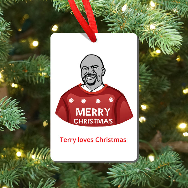 A Christmas ornament hanging from a red ribbon, featuring an illustration of Brooklyn Nine Nine's Terry wearing a red sweater with snowflakes and the words 'MERRY CHRISTMAS'. Below the illustration, the text reads 'Terry loves Christmas'. shown against a blurred Christmas tree background with lights.