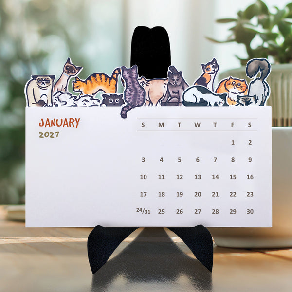 A variety of illustrated cats on the top of each die cut month on a desktop calendar with a black easel sitting on a table.