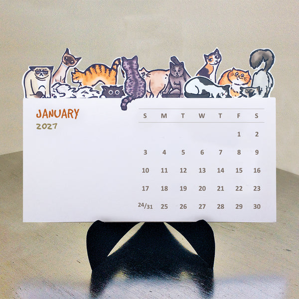 A variety of illustrated cats on the top of each die cut month on a desktop calendar with a black easel sitting on a table.