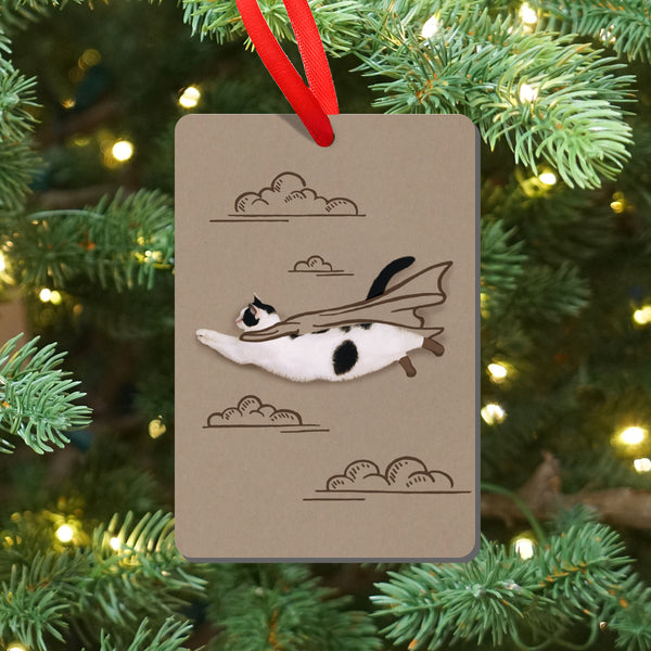 Decorative ornament with a flying cat on a beige magnet, hanging on a Christmas tree.