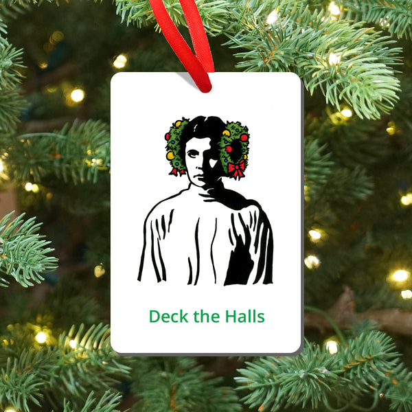 A white rectangular Christmas ornament hanging from a red ribbon, featuring a black-and-white illustrated portrait of Princess Leia with wreaths on the side of her head and the green text 'Deck the Halls' at the bottom. The ornament is photographed against a blurred Christmas tree with green needles and warm string lights.