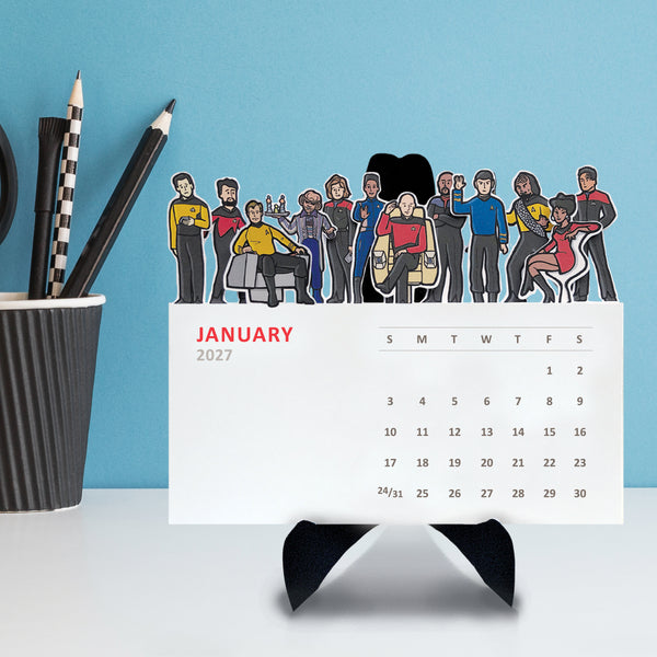 Illustrations of a variety of Star Trek characters on top of each monthly card on a desktop calendar on a black easel sitting on a table.