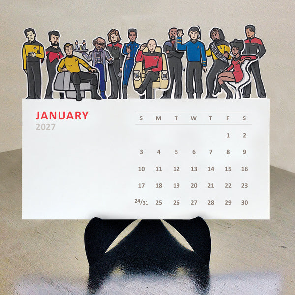 Illustrations of a variety of Star Trek characters on top of each monthly card on a desktop calendar on a black easel sitting on a table.
