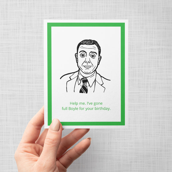 A hand holding a novelty birthday card featuring a black line-art portrait of Brooklyn Nine Nine's Boyle in a suit and tie, with green text at the bottom reading, 'Help me. I've gone full Boyle for your birthday.'