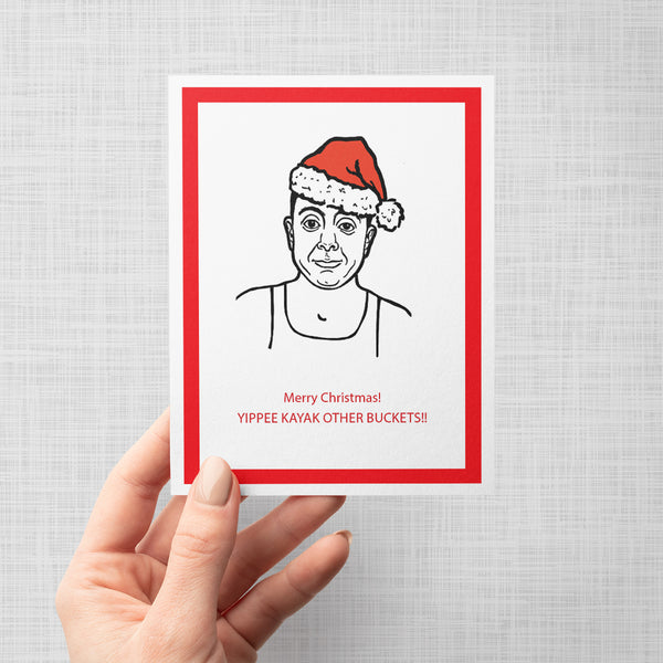 A hand holding a Christmas greeting card featuring a black-and-white line drawing of Brooklyn Nine Nine's Boyle wearing a red Santa hat, with red text reading 'Merry Christmas! YIPPEE KAYAK OTHER BUCKETS!!' on a white card with a red border, shown against a light gray background.
