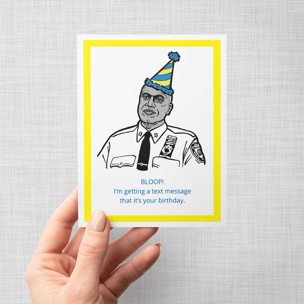 A hand holding a novelty greeting card featuring an illustration of a Brooklyn Nine Nine's Captain Holt wearing a blue and yellow striped party hat. The card has a white background with a thick yellow border and blue text that reads, 'BLOOP! I'm getting a text message that it's your birthday.