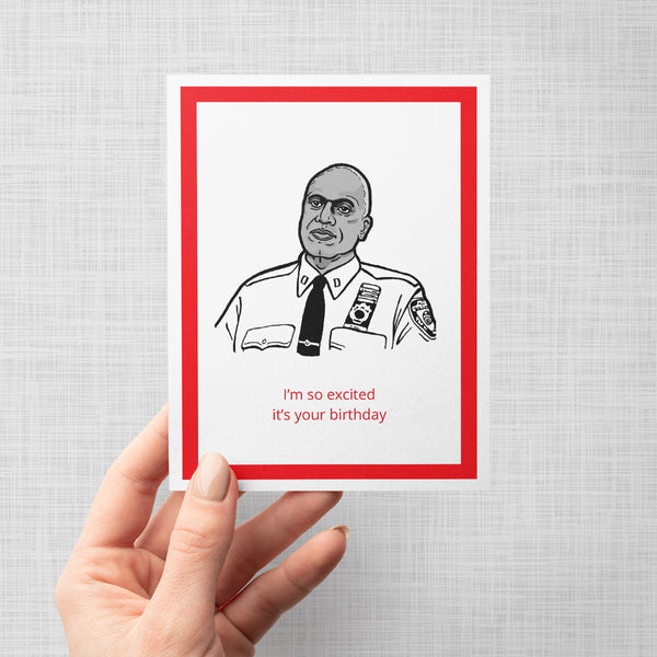 A hand holding a birthday greeting card featuring a black and white illustration of Brooklyn Nine Nine's Captain Holt with ra ed border and the text 'I'm so excited it's your birthday' in red on a white card, shown against a light gray background.