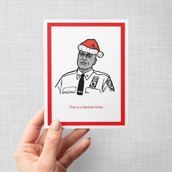 A hand holding a greeting card featuring a black and white illustration of Brooklyn Nine Nine's Captain Holt wearing a red Santa hat, with the red text 'This is a festive time.' 
