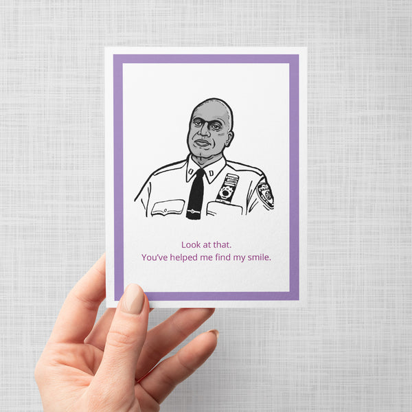 A hand holding a greeting card featuring a black and white line art illustration of Brooklyn Nine Nine's Captain Holt with the text 'Look at that. You've helped me find my smile.'