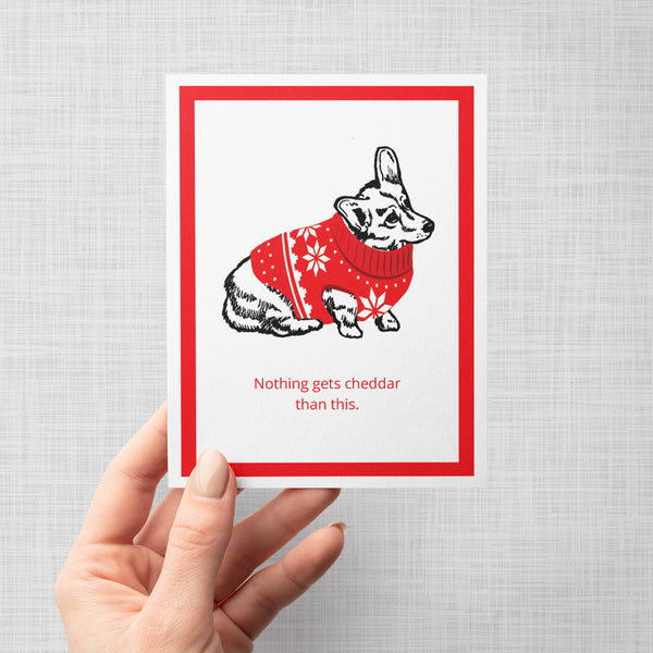 A hand holds a white greeting card with a red border, featuring Brooklyn Nine Nine's cheddar dog wearing a red holiday sweater with white snowflake patterns. The card has the text 'Nothing gets cheddar than this.