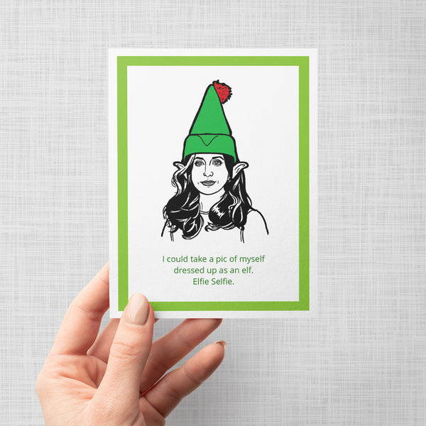 A hand holding a greeting card featuring a black and white line art illustration of Brooklyn Nine Nine's Gina wearing a green elf hat with a red pom-pom, framed by a green border on a white background. Below the illustration, green text reads: 'I could take a pic of myself dressed up as an elf. Elfie Selfie.'