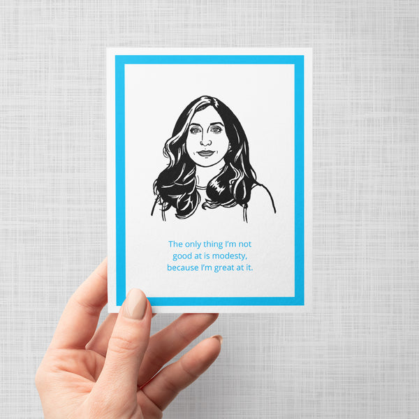 A hand holding a greeting card featuring a black-and-white sketch of Brooklyn Nine Nine's Gina with the quote, 'The only thing I'm not good at is modesty, because I'm great at it.' in blue text, framed by a bright blue border on a white background, shown against a light gray backdrop.