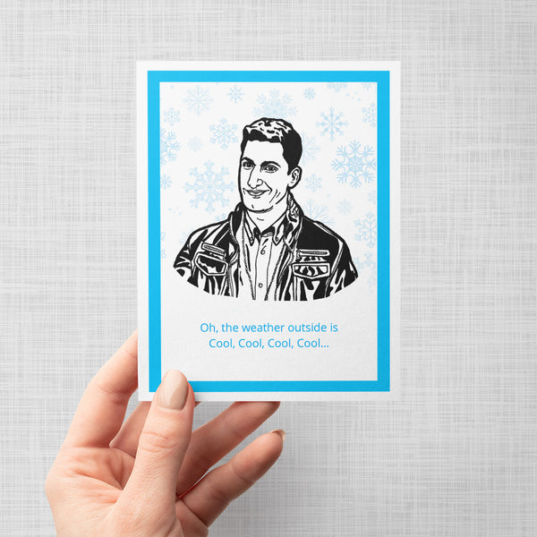 A hand holding a greeting card featuring a black-and-white illustrated portrait of Brooklyn Nine Nine's Jake Peralta, surrounded by light blue snowflakes and ending with the text 'Oh, the weather outside is Cool, Cool, Cool, Cool.'