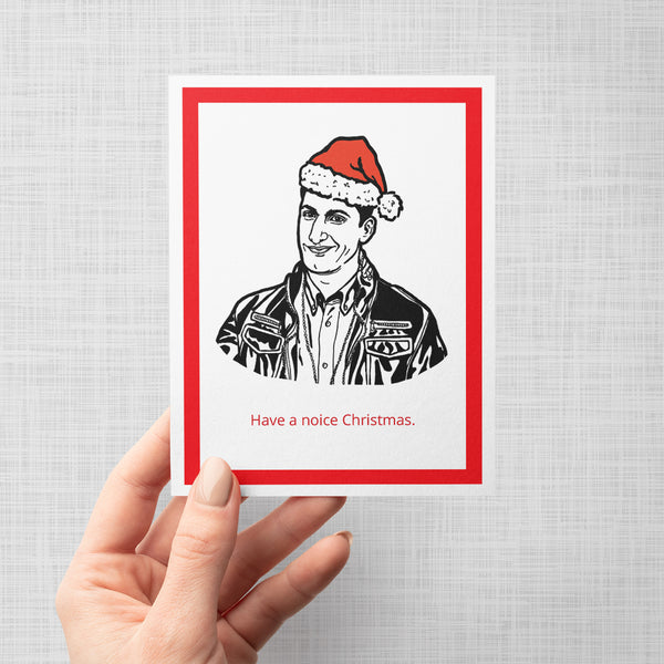 A hand holding a greeting card featuring a black and white line art illustration of Brooklyn Nine Nine's Jake Peralts wearing a red Santa hat, with the text 'Have a noice Christmas.' printed below in red.