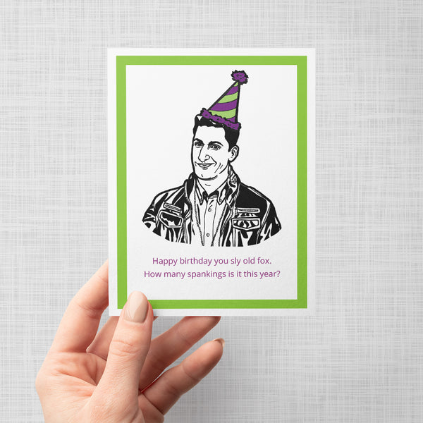 A hand holding a greeting card featuring a black and white line art illustration of Brooklyn Nine Nine's Jake Peralta wearing a purple and green striped party hat, with a bright green border and pink text at the bottom that reads, 'Happy birthday you sly old fox. How many spankings is it this year?' The card is shown against a neutral gray background.