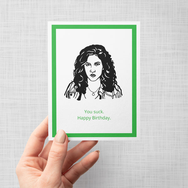A hand holding a greeting card featuring a black and white line art portrait of Brooklyn Nine Nine's Rosa bordered by a green inner frame and a white outer border. Below the portrait, the text reads 'You suck. Happy Birthday.'