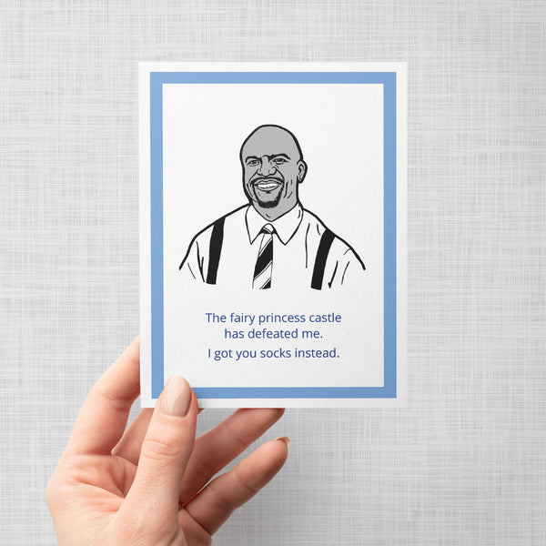 A hand holding a greeting card featuring a black and white line art illustration of a Brooklyn Nine Nine's Terry with blue text below reading, 'The fairy princess castle has defeated me. I got you socks instead.'
