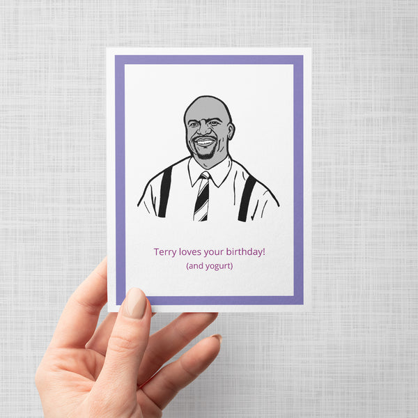A hand holding a novelty greeting card featuring a black and white line art illustration of Brooklyn Nine Nine's Terry Fox, with purple text at the bottom reading 'Terry loves your birthday! (and yogurt)'. The card is presented on a neutral gray background.