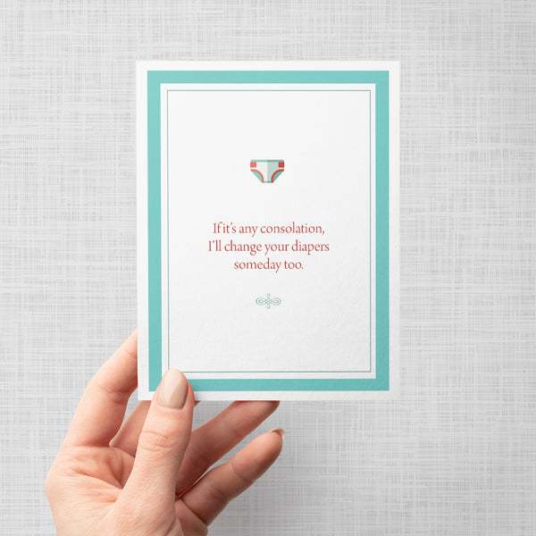 Hand holding greeting card with a cartoon diaper and humorous text 'If it's any consolation, I'll change your diapers someday too' on a gray background