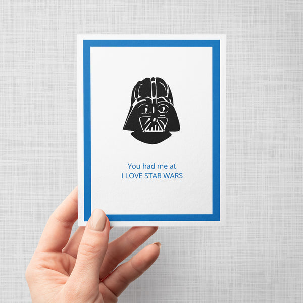 A hand holding a greeting card featuring a black illustration of Darth Vader above blue text that reads 'You had me at I LOVE STAR WARS', set within a blue border on a white background, shown against a light gray backdrop.