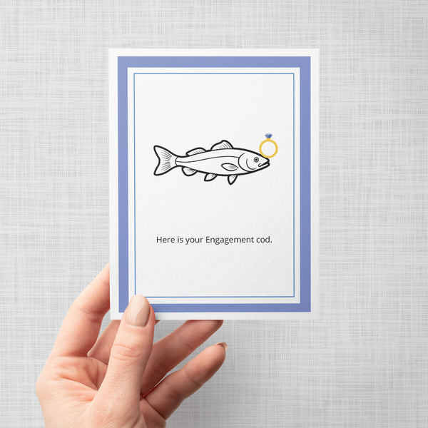 Greeting card with a fish illustration holding an engagement ring and text 'Here is your Engagement cod' being held by a hand