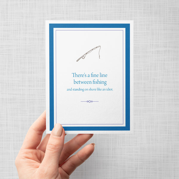 Hand holding greeting card with fishing pole illustration and text 'There's a fine line between fishing and standing on shore like an idiot'