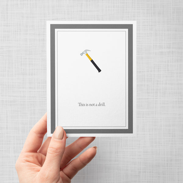 Hand holding greeting card with a hammer illustration and text 'This is not a drill' on a gray background
