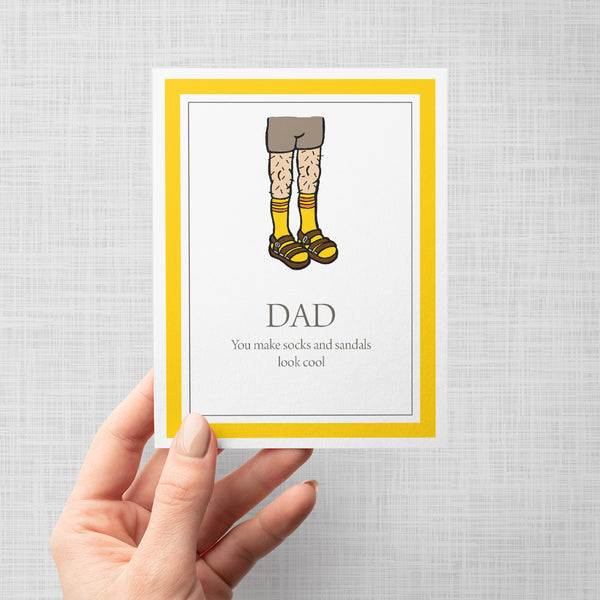 Hand holding greeting card with illustration of socks and sandals with text 'Dad you make socks and sandals look cool' on a gray background