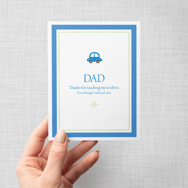 Hand holding greeting card with a blue car illustration and text 'Dad thanks for teaching me to drive even though I still suck at it' on a gray background