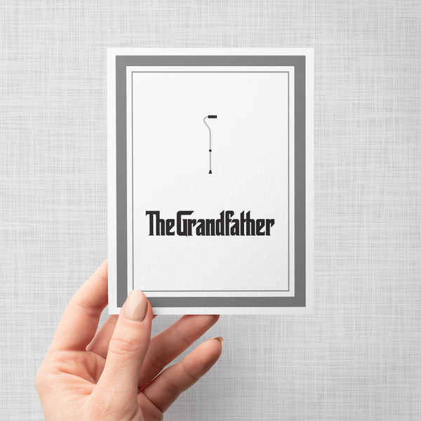 Hand holding greeting card with cane illustration and text 'The Grandfather' on a gray background