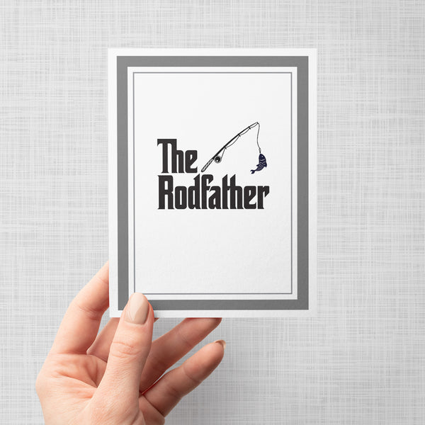 Hand holding greeting card with fishing pole and fish illustration and text 'The Rodfather' on a gray background