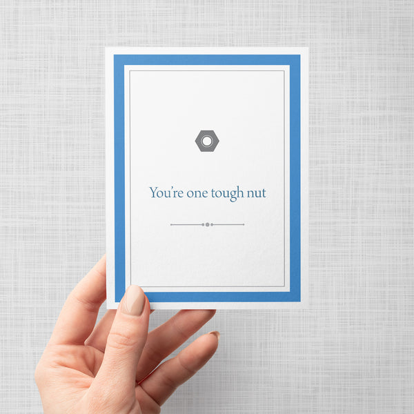 Hand holding greeting card with metal nut illustration and text 'You're one tough nut' on a gray background