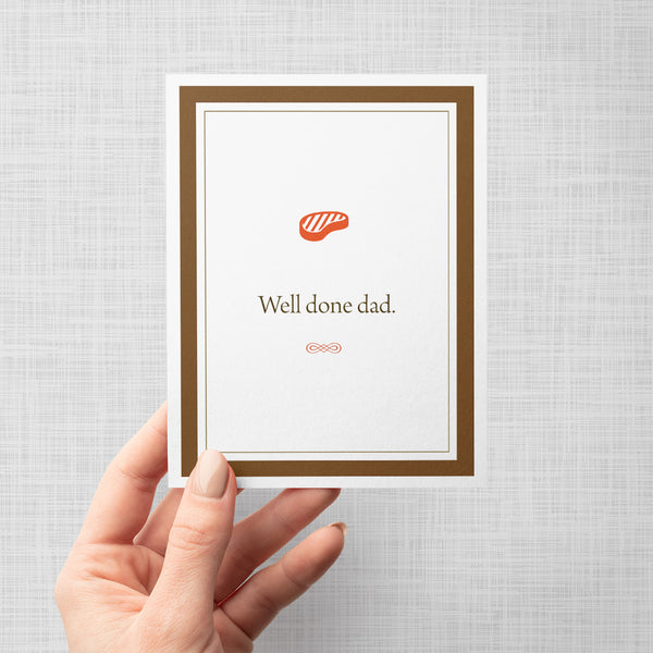 Hand holding a greeting card with a red steak icon and text 'Well done dad' on a gray background