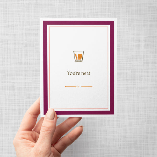 Hand holding greeting card with a glass of scotch graphic and text 'You're neat' on a gray background