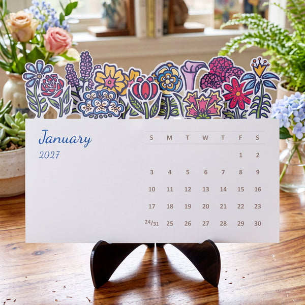 A decorative 2027 desk calendar page with colorful floral illustrations across the top, displayed on a black stand on a wooden table with flowers in the background.