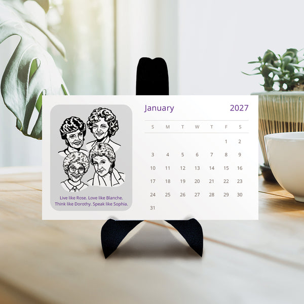 A white monthly 2027 calendar with black and white illustration of the Golden Girls characters displayed on a black stand against a beige wall and floor. 