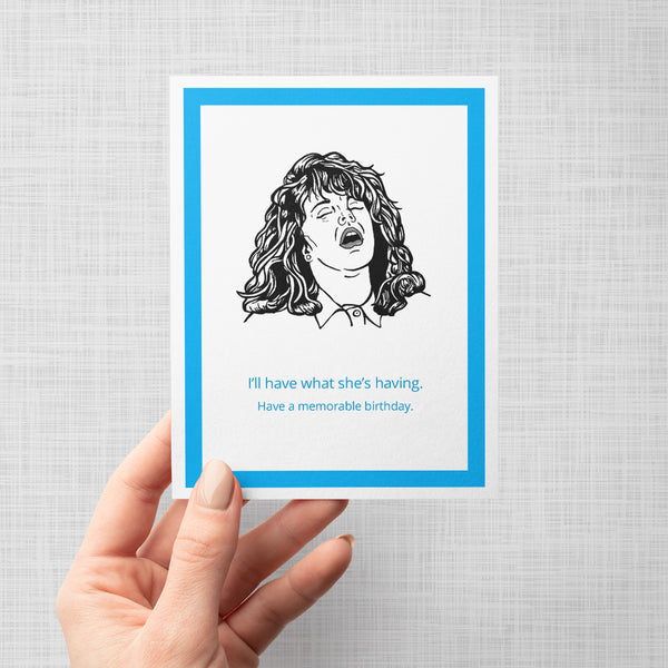 Hand holding Illustration of a greeting card with Sally faking an orgasm from the movie When Harry Met Sally. Text says 'I'll have what she's having. Have a memorable birthday'