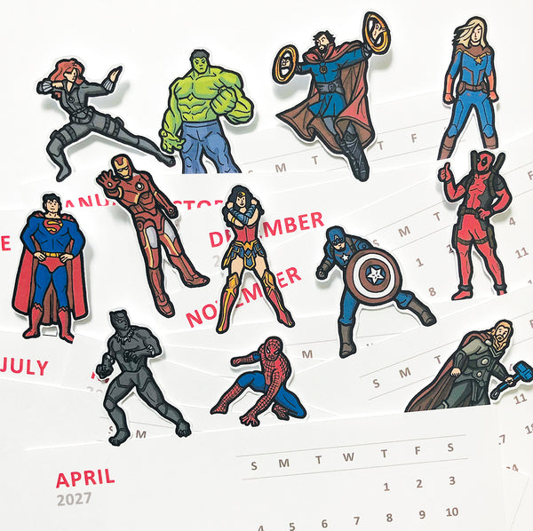 A set of die-cut 2027 calendar cards featuring Marvel superheroes arranged on top of a white calendar page. The characters include Black Widow, Hulk, Captain America, Thor, Scarlet Witch, Captain Marvel, Superman, Iron Man, Wonder Woman, Deadpool, Black Panther, Spider-Man, and Thor.