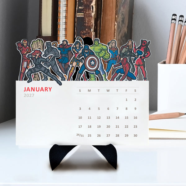 A 2027 desk calendar featuring a colorful row of Marvel and DC superhero character illustrations, displayed on a light colored table.