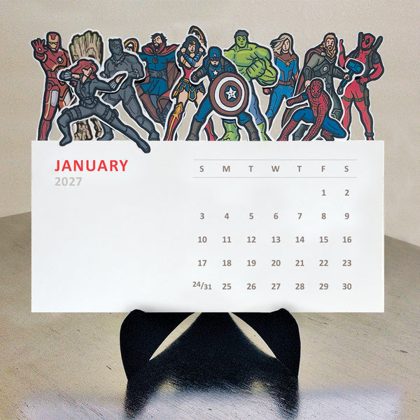 A 2027 desk calendar featuring a colorful row of Marvel and DC superhero character illustrations, displayed on a black stand against a light beige wall.