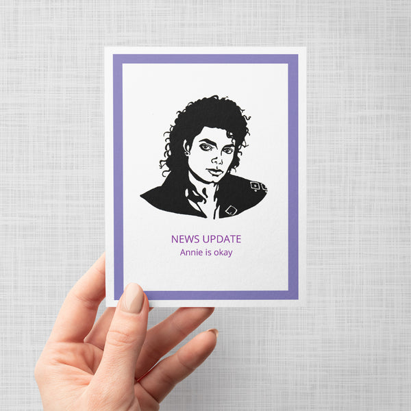 Hand holding a card with a black and white portrait of Michael Jackson and 'NEWS UPDATE Annie is okay' text on a gray background
