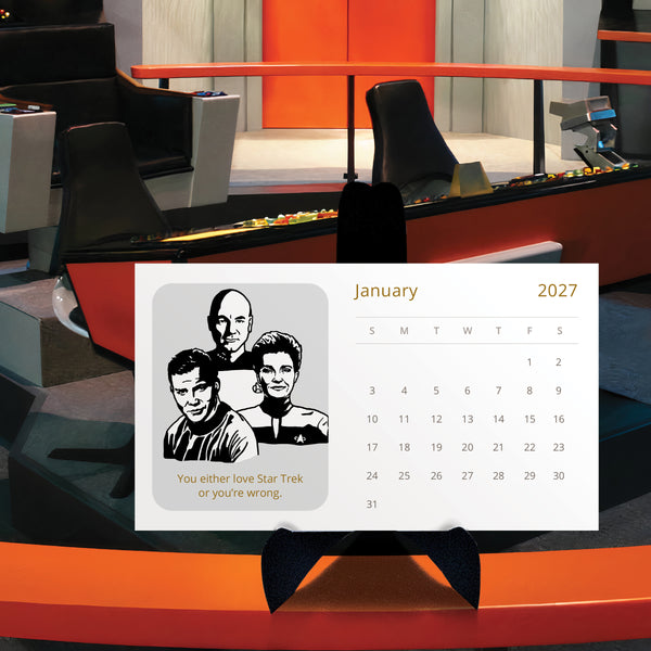 A 2027 Star Trek desk calendar featuring a black-and-white Star Trek captains illustration and the phrase 'You either love Star Trek or your wrong,' displayed on a black stand on the rail of the bridge of the Enterprise.
