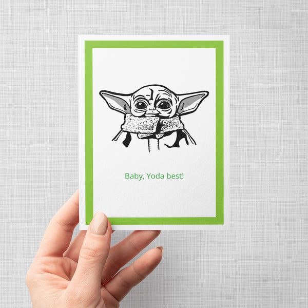 A hand holding a greeting card featuring a black line art illustration of Baby Yoda from Star Wars with the text 'Baby, Yoda best!' in green, framed by a bright green border on a white card against a light gray background.