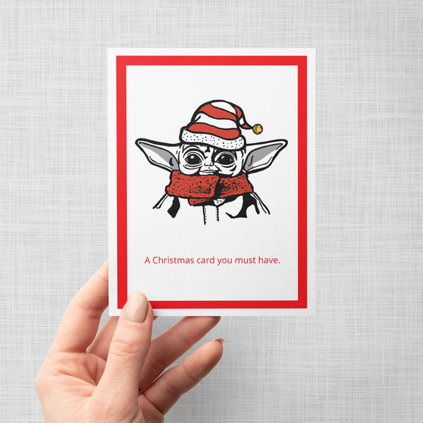 A hand holding a Christmas-themed greeting card featuring a cartoon illustration of Grogu wearing a red and white Santa hat and matching red scarf, with the text 'A Christmas card you must have.' printed below.