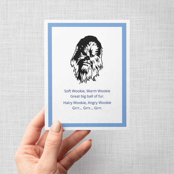 A hand holding a greeting card featuring a black line-art illustration of Chewbacca above blue text that reads, 'Soft Wookie, Warm Wookie Great big ball of fur. Hairy Wookie, Angry Wookie Grrr Grrr Grrr' on a light grey background.
